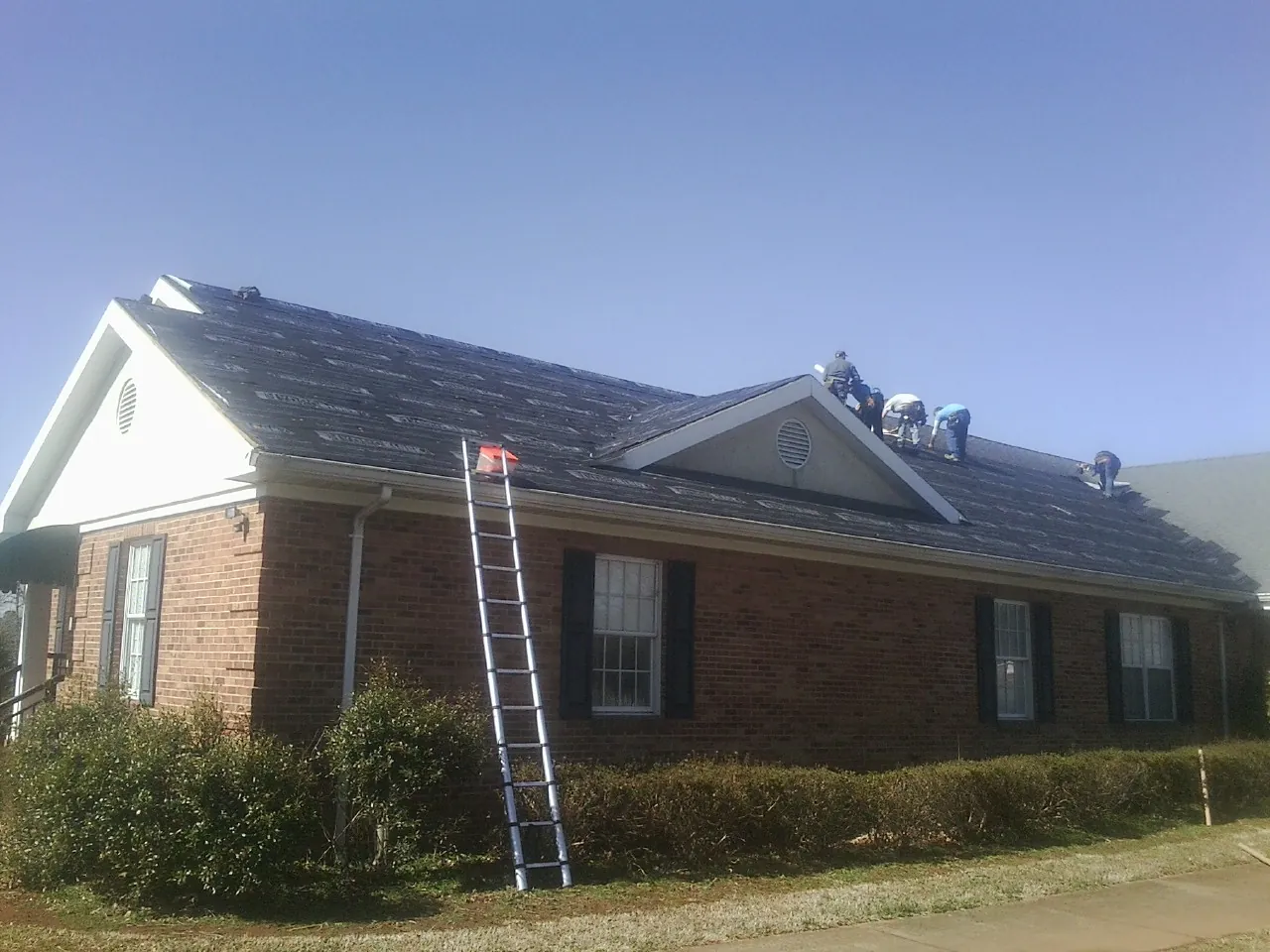 Crew tearing shingles off a brick ranch roof with a ladder set — Emergency Roof Repair work by our crew in Tecumseh